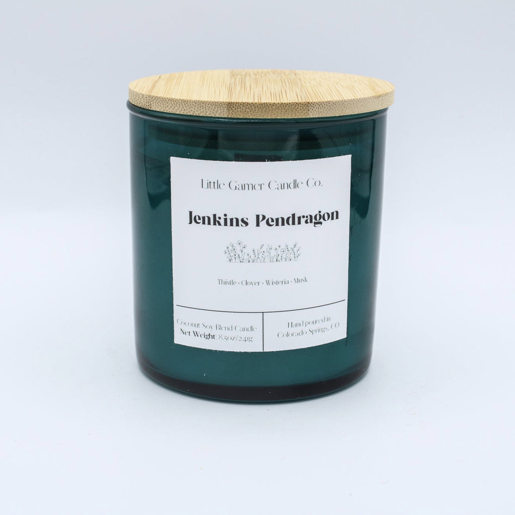 A dark green glass jar candle with a light bamboo wood lid and a white paper label reading 'Little Gassy Candle Co. jenkins Pendragon'.