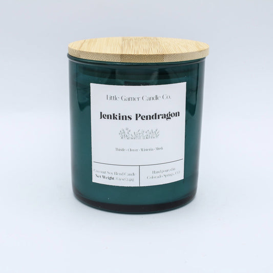 A dark green glass jar candle with a light bamboo wood lid and a white paper label reading 'Little Gassy Candle Co. jenkins Pendragon'.