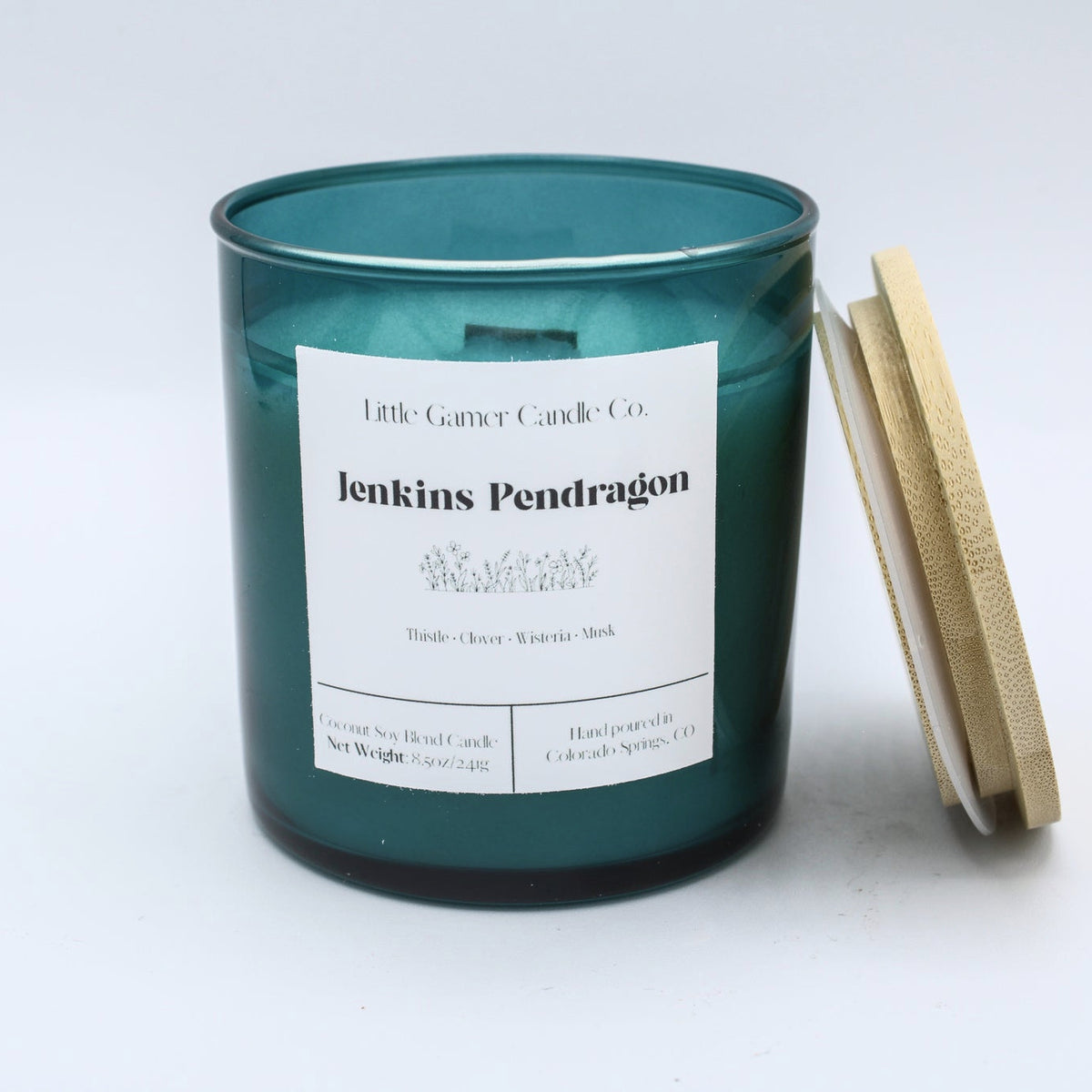 A teal glass jar candle with a white paper label reading 'Little Century Candle Co. Jenkins Pendragon' and a light wood lid leaning against the right side, photographed on a clean white background.