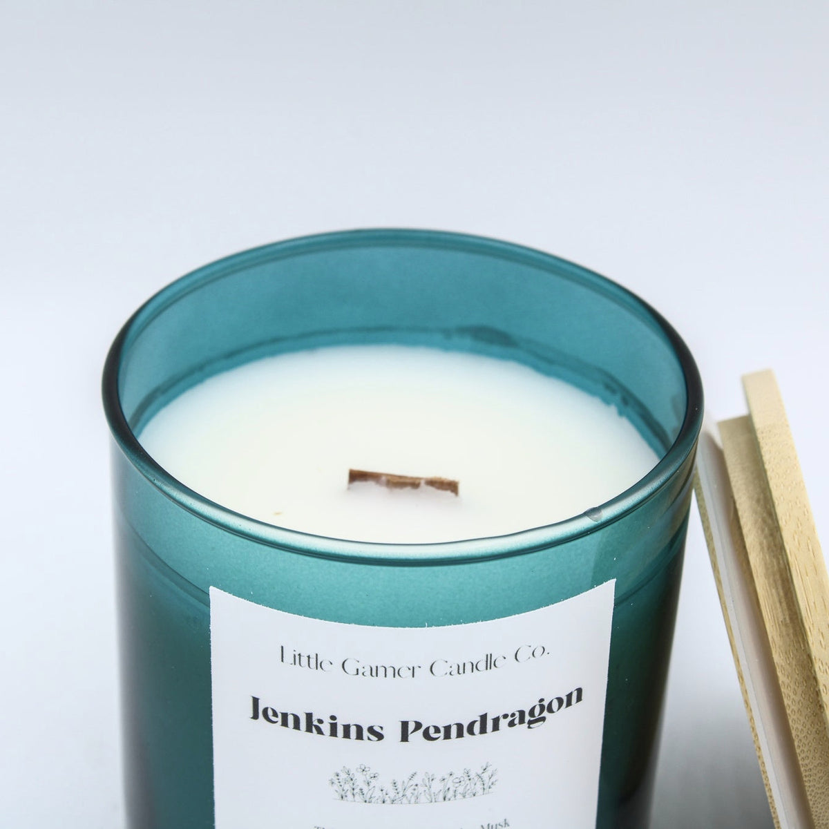 A teal glass jar candle with white wax and a wooden wick, labeled 'Jenkins Pendragon' by Little Giant Candle Co., shown with a gold-colored lid resting beside it on a light gray background.