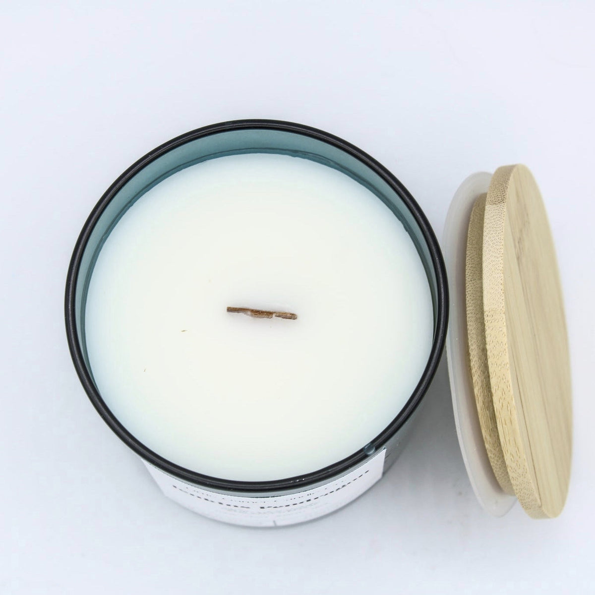 A top-down view of a white wax candle in a black-rimmed jar with a light wood lid resting in front, on a clean white background.