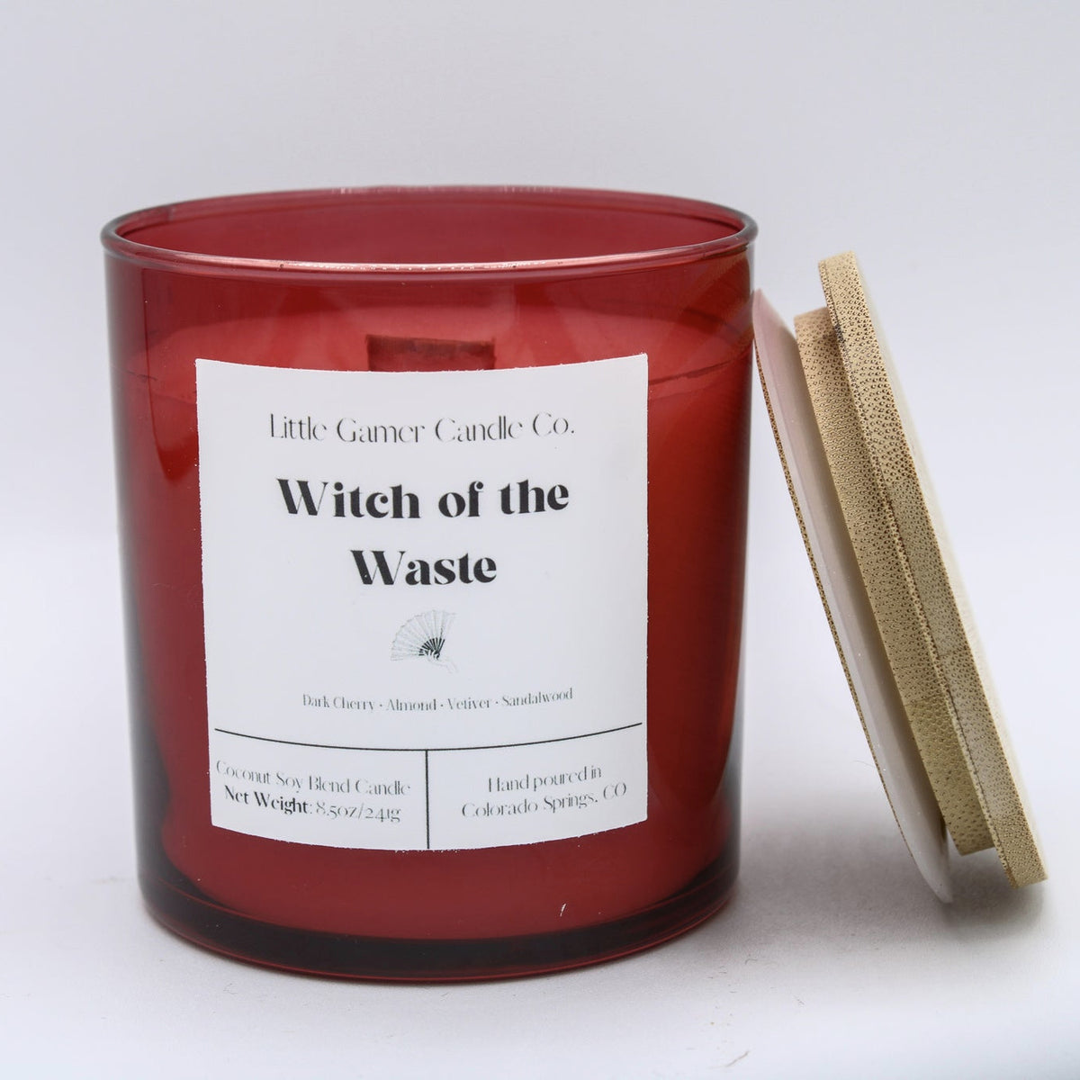 A red glass jar candle with a white label reading 'Little Gamer Candle Co. Witch of the Waste,' shown next to its light wood lid on a white background.