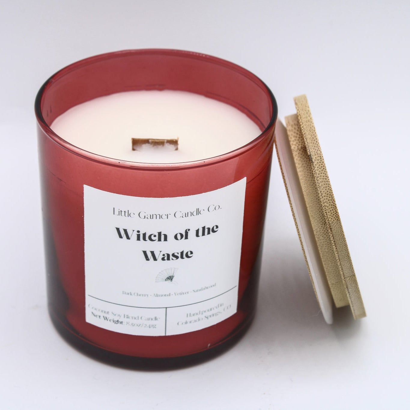 A red glass jar candle with white wax and a white label reading 'Witch of the Waste' by Little Gassy Candle Co, shown on a white background with its wooden lid leaning against it.