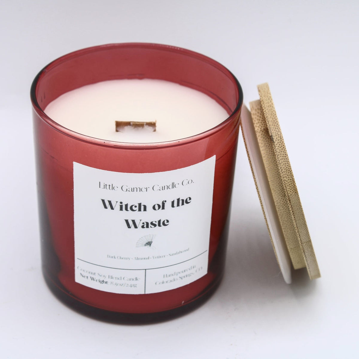 A red glass jar candle with white wax and a white label reading 'Witch of the Waste' by Little Gassy Candle Co, shown on a white background with its wooden lid leaning against it.