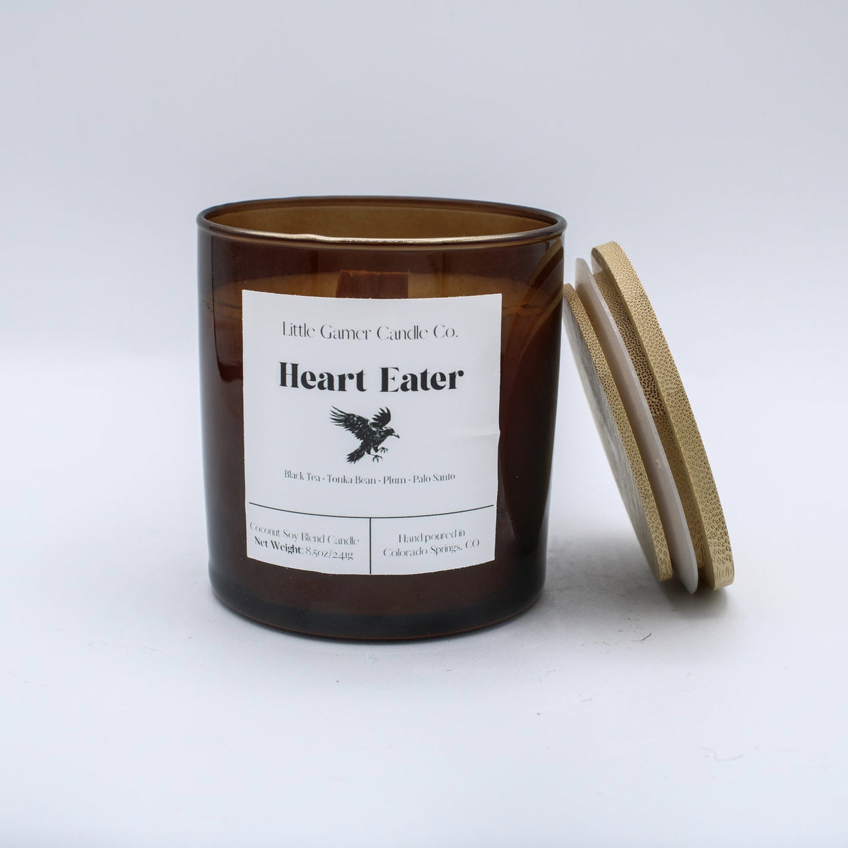 An amber glass jar candle with a white label reading 'Little Goose Candles Co. Heart Eater' and a light wood lid leaning against the right side, photographed on a clean white background.