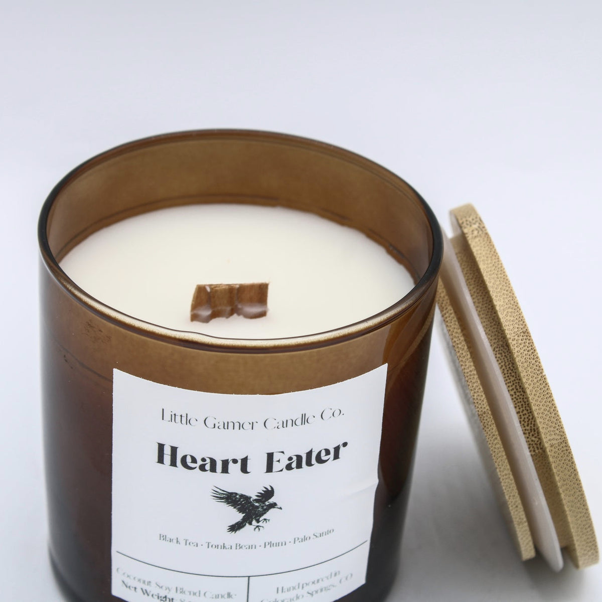 An amber glass jar candle with white wax and a wooden wick, labeled 'Heart Eater' by Little Gather Candle Co., shown with its bamboo lid leaning against it on a white background.