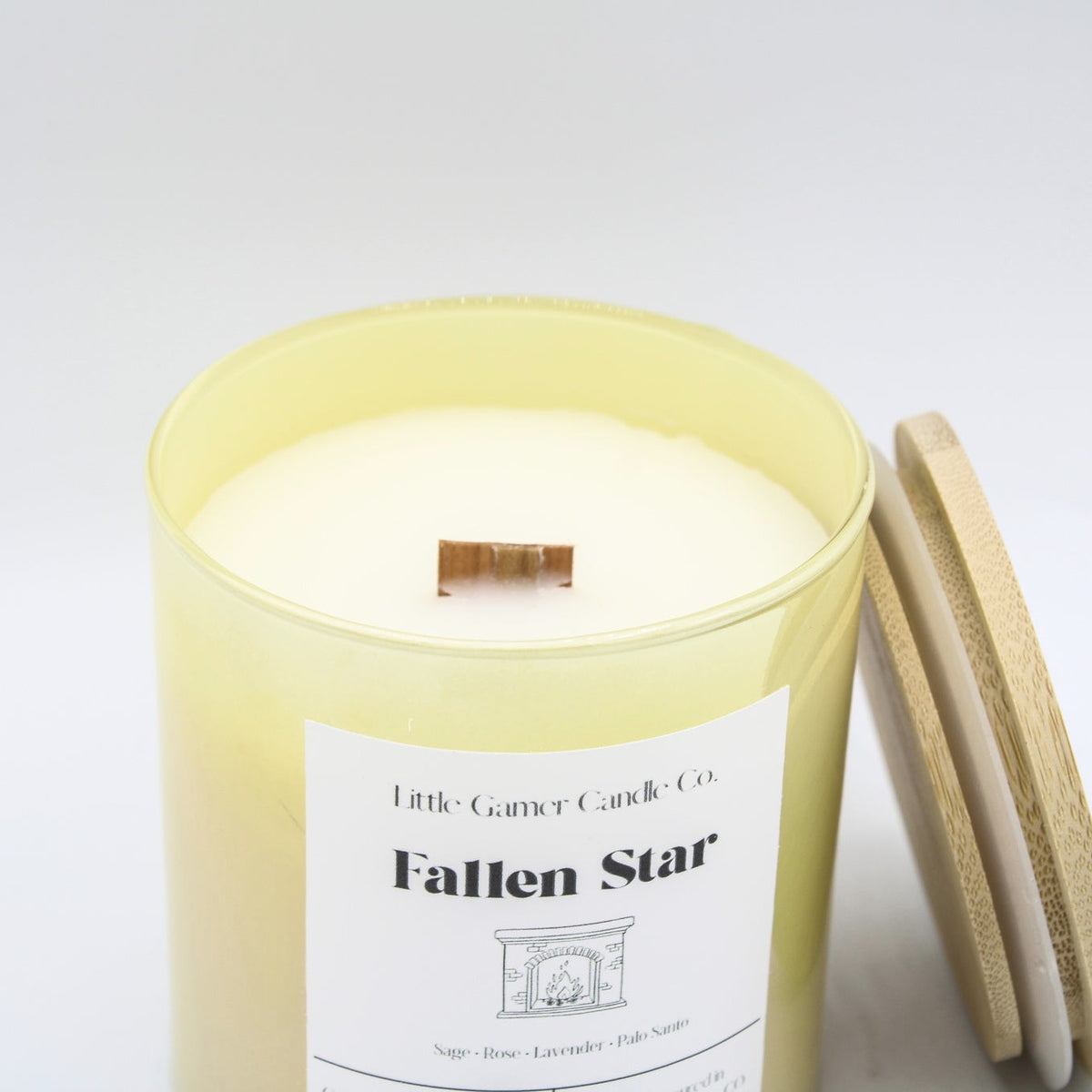 A close up of a  yellow iridescent jar with a white label reading "Little Gamer Candle Co. Fallen Star" with a light bamboo lid leaning against it on a white background.