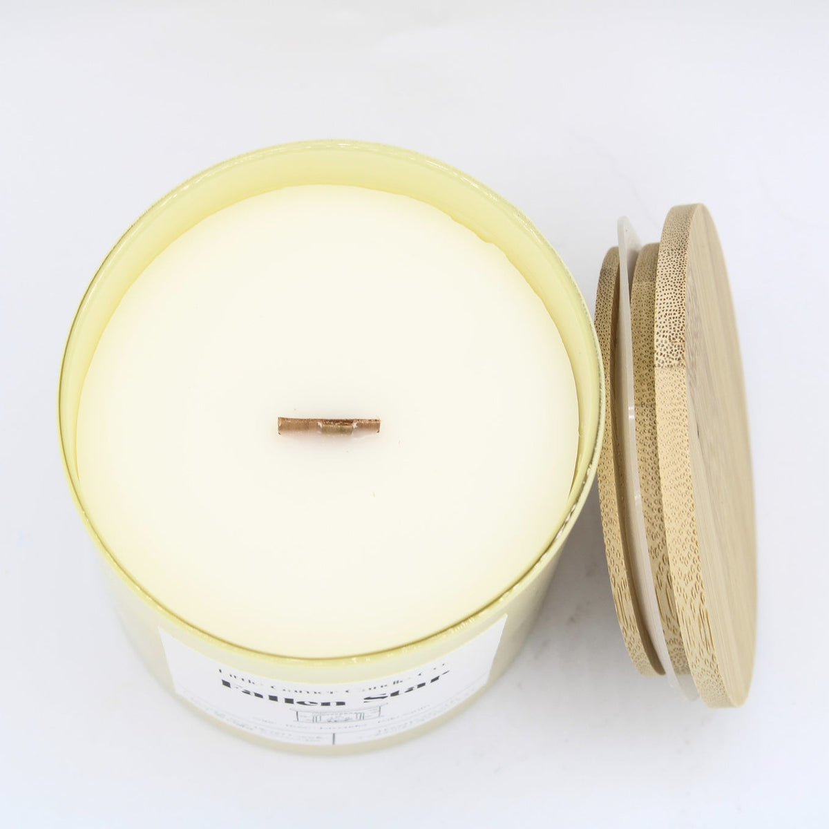 A soy wax candle in a clear glass jar with a pale yellow tint, featuring a single wick and shown next to its bamboo lid on a white background.