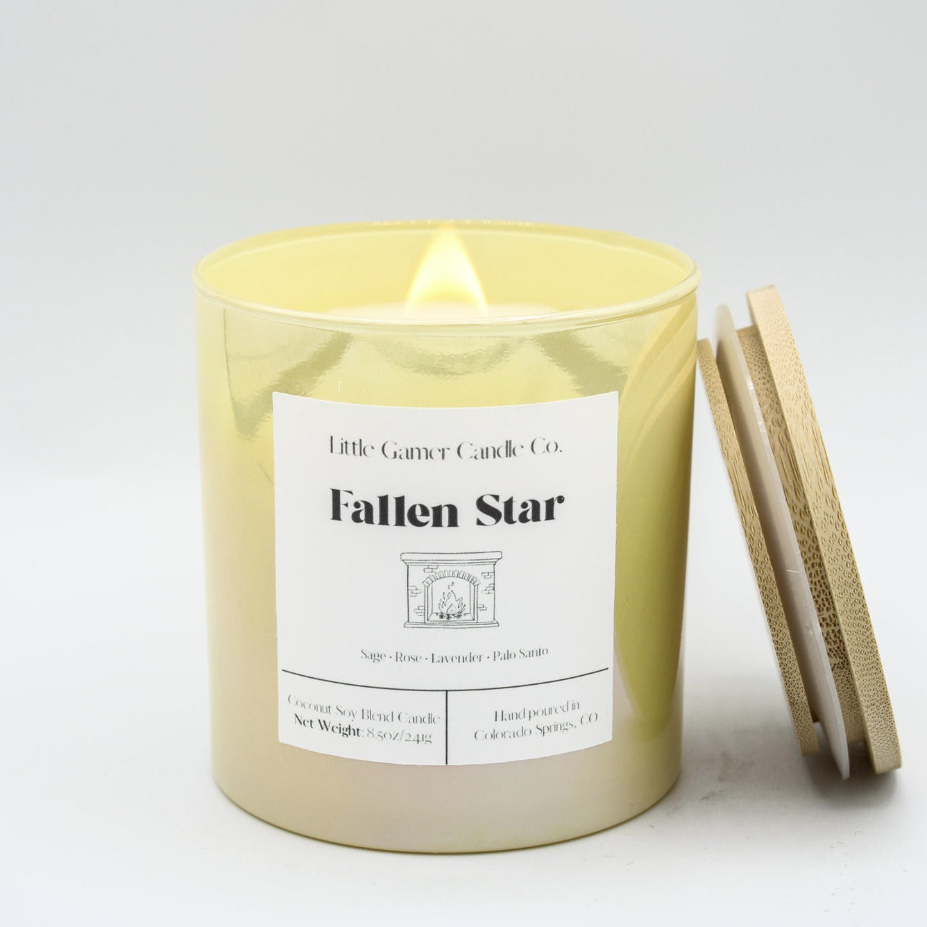 A lit candle in a yellow iridescent jar with a white label reading "Little Gamer Candle Co. Fallen Star" with a light bamboo lid, leaning against the jar on a white background.