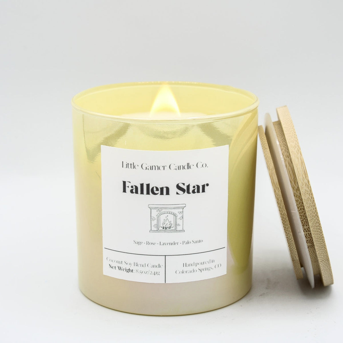 A lit candle in a yellow iridescent jar with a white label reading "Little Gamer Candle Co. Fallen Star" with a light bamboo lid, leaning against the jar on a white background.