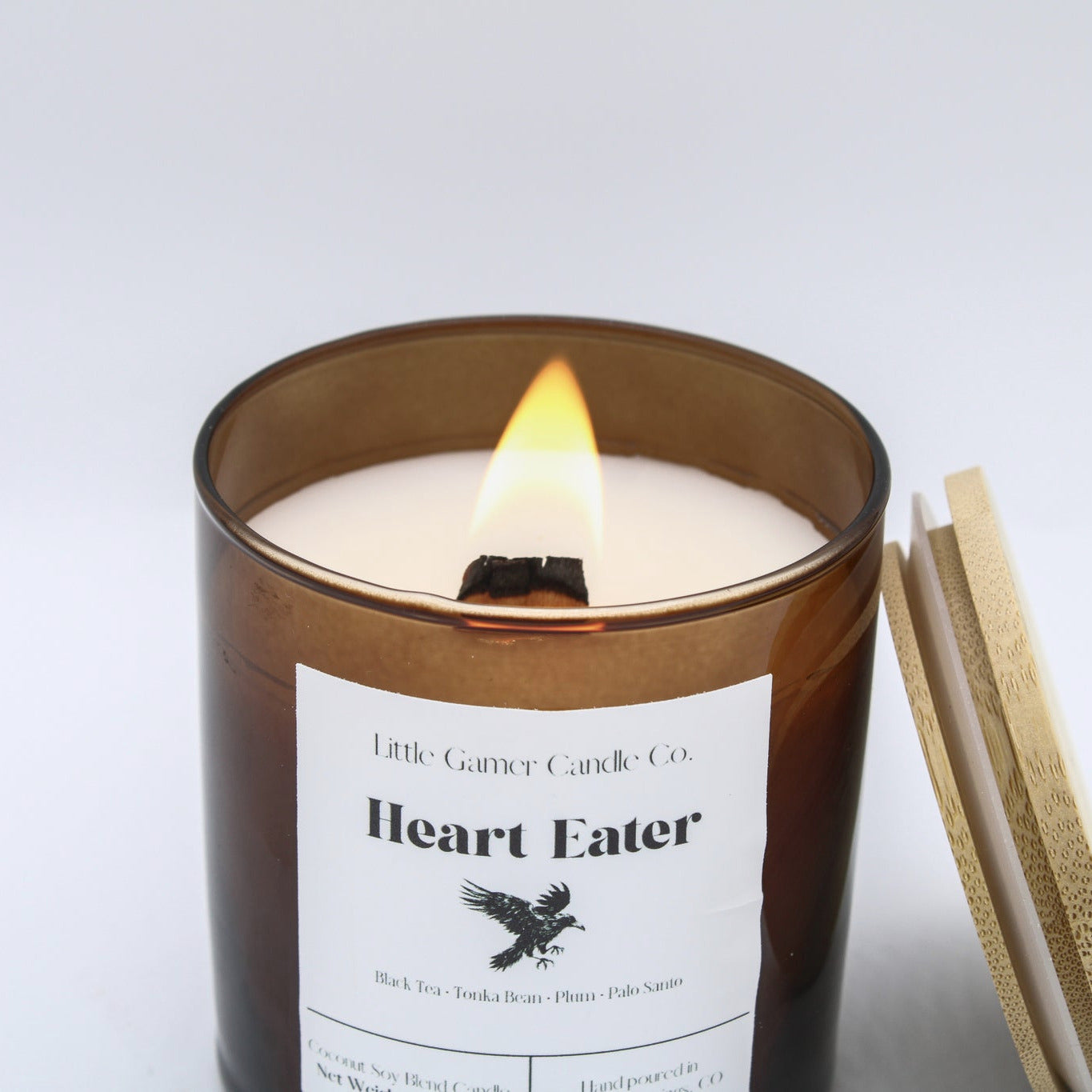 An amber glass jar candle with a lit wick and visible flame, labeled 'Heart Eater' by Little Gander Candle Co, shown on a clean white background with a gold-toned matchbook or lid leaning against the right side.