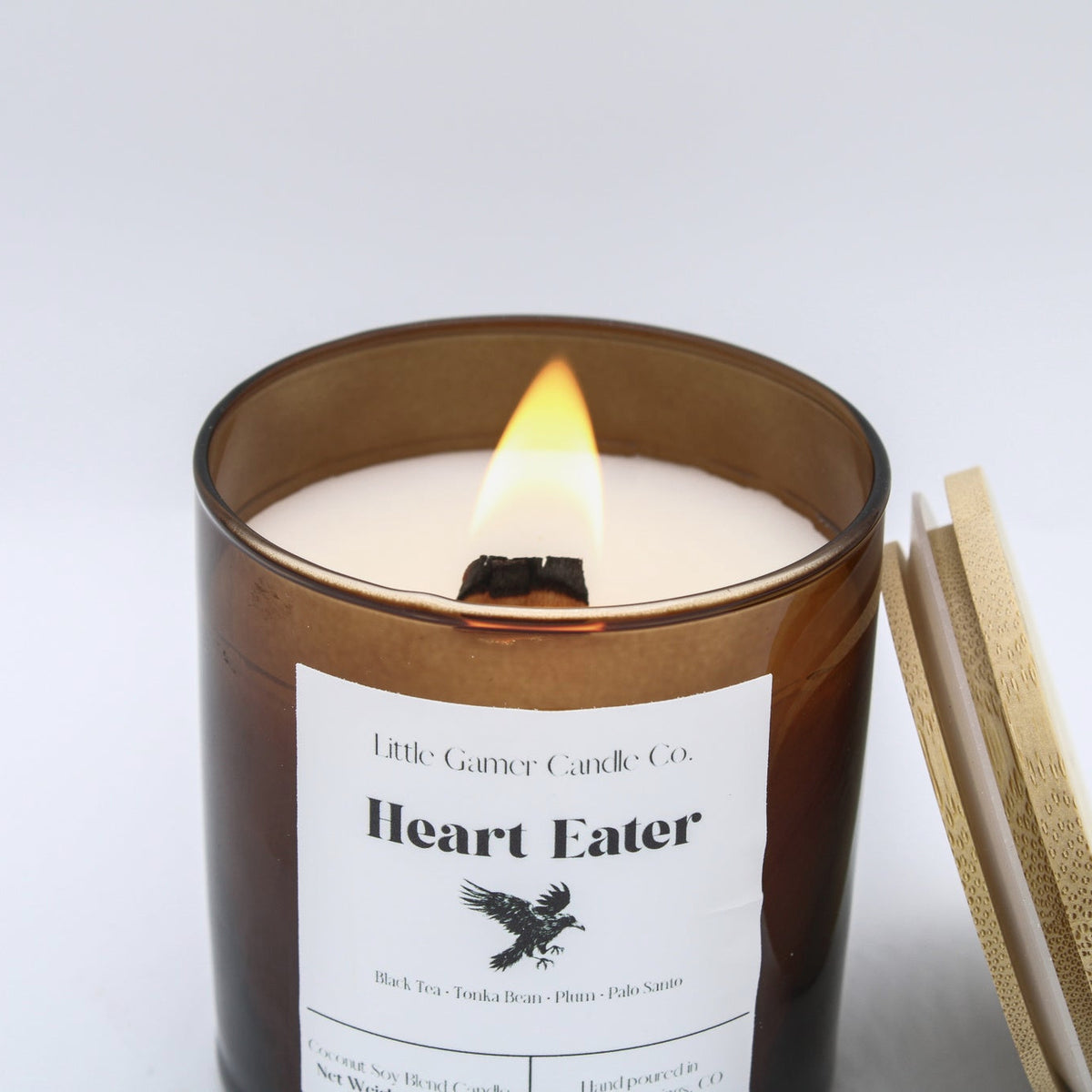 An amber glass jar candle with a lit wick and visible flame, labeled 'Heart Eater' by Little Gander Candle Co, shown on a clean white background with a gold-toned matchbook or lid leaning against the right side.