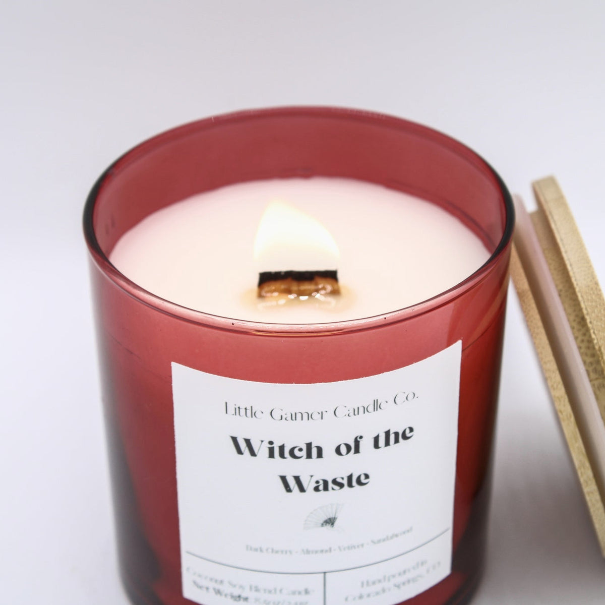 A red glass jar candle with a white label reading 'Little Gander Candle Co. Witch of the Waste' and a lit wick, shown on a white background with a wooden lid leaning against the jar.