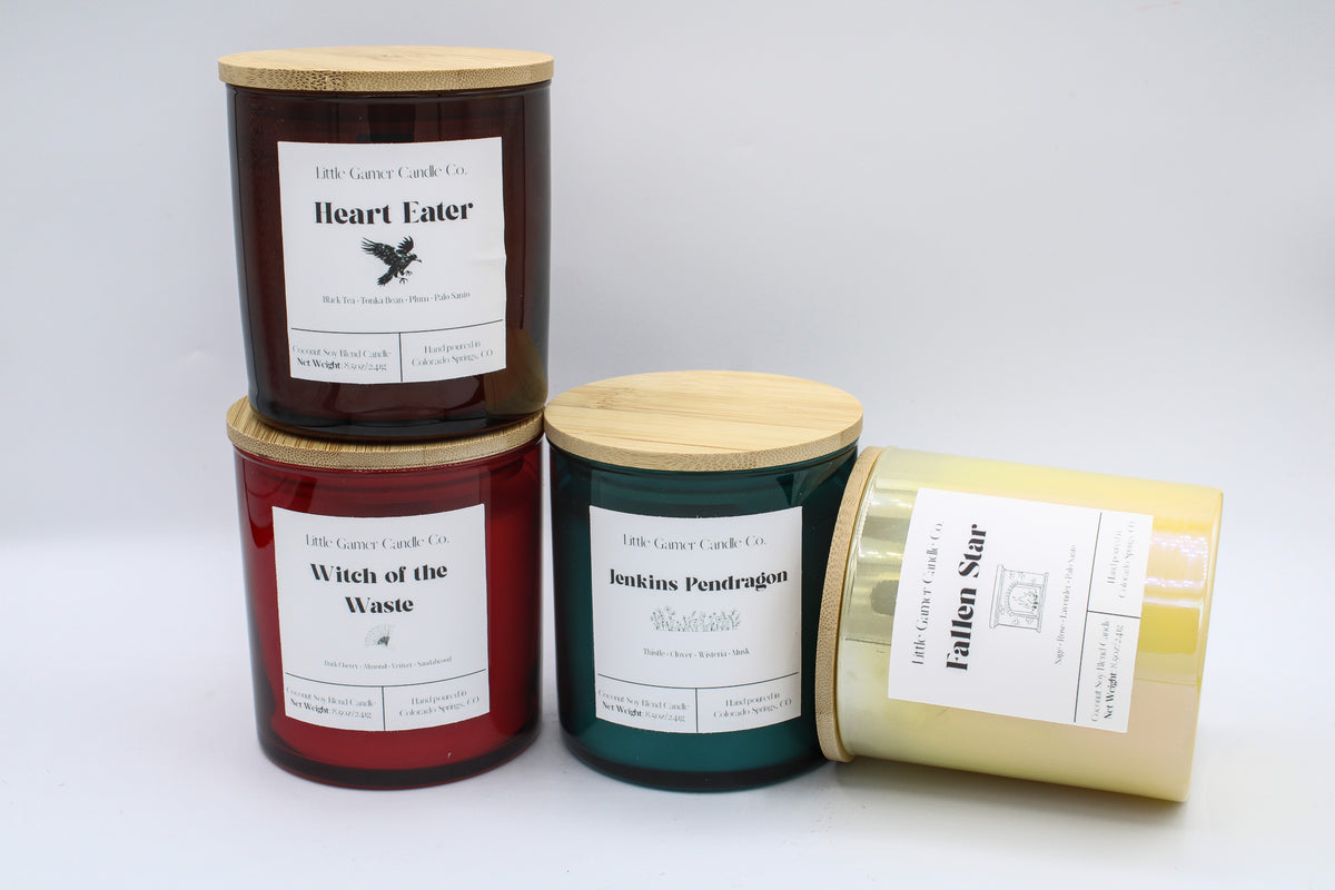 Four candles in glass jars with bamboo lids, arranged on a light gray background. The labels read 'coconut soy blend candles' and include 'Heart Eater', 'Witch of the Waste', 'Jenkins Pendragon', and 'Fallen Star'.