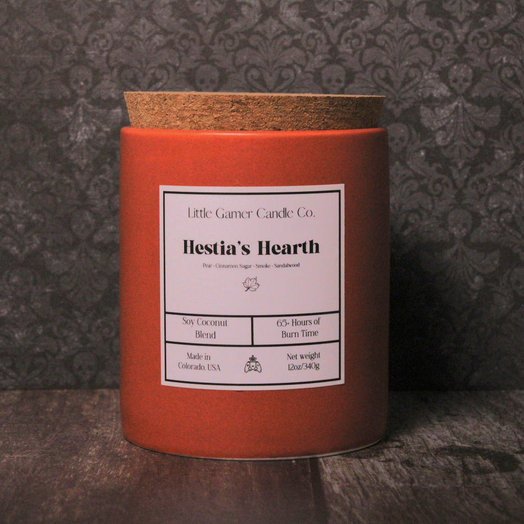 A red cylindrical candle tin with a cork lid and a white label reading 'Little Garner Candle Co. Hestia's Hearth,' set on a dark wooden surface against a dark patterned wallpaper background.