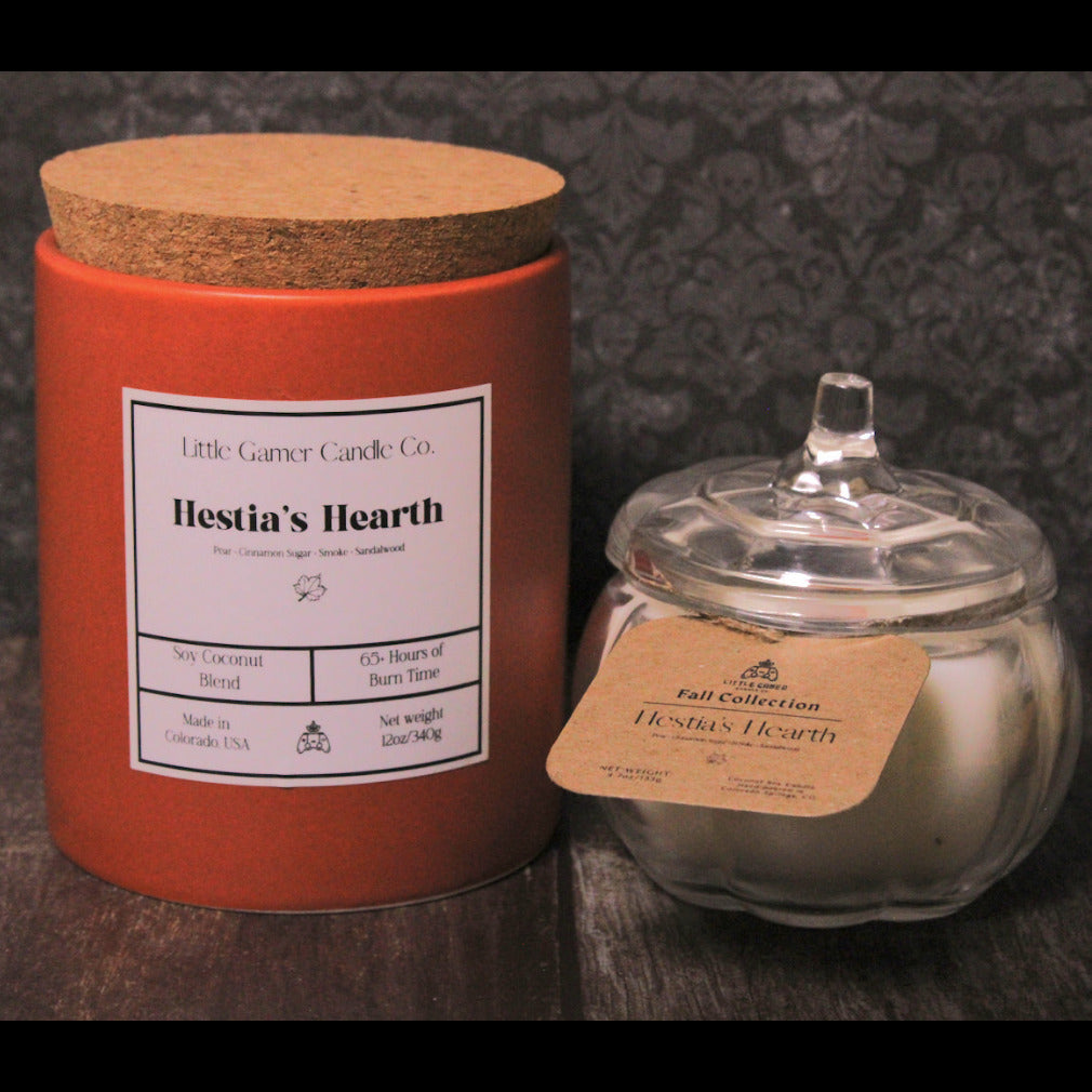 A red cylindrical candle tin with a cork lid stands next to a small clear glass jar candle with a glass lid and kraft paper label. Both containers show the Little Gamer Candle Co.