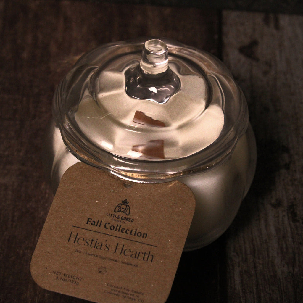 A glass jar candle with a lid and a kraft paper label reading 'Fall Collection Hestia's Hearth' on a dark rustic wooden surface.