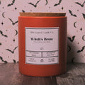 A red-orange candle jar with a cork lid and a white label reading 'Little Gamer Candle Co. Witch's Brew,' set on a dark wooden surface against a pink patterned backdrop with black bats and moons.