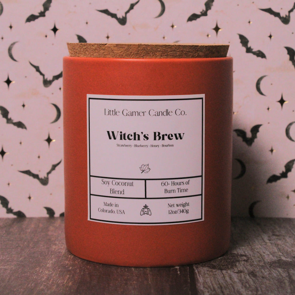 A red-orange candle jar with a cork lid and a white label reading 'Little Gamer Candle Co. Witch's Brew,' set on a dark wooden surface against a pink patterned backdrop with black bats and moons.