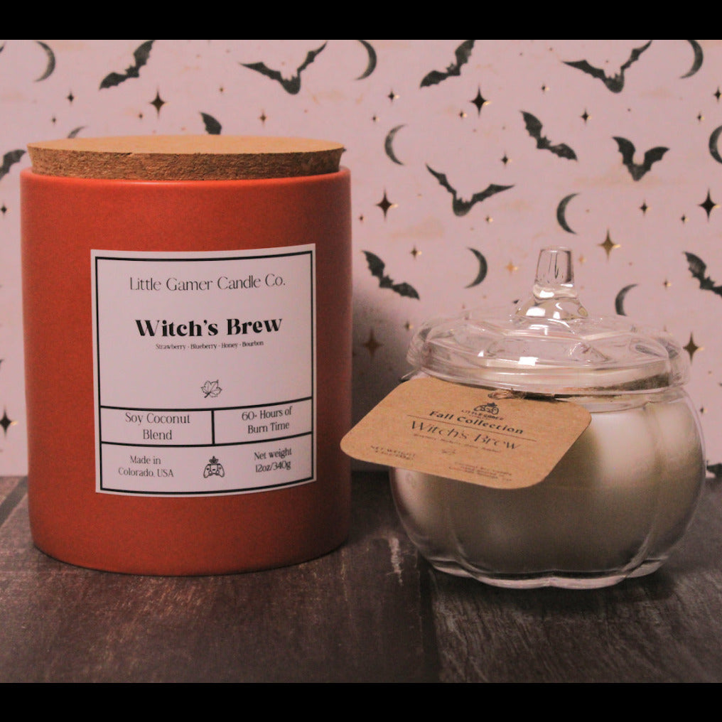 A red cylindrical candle tin labeled 'Witch's Brew' by Little Gamer Candle Co. stands next to an open clear glass jar candle with pale wax and a kraft paper tag, set on a dark tabletop against a pink patterned backdrop with black bat and moon motifs.