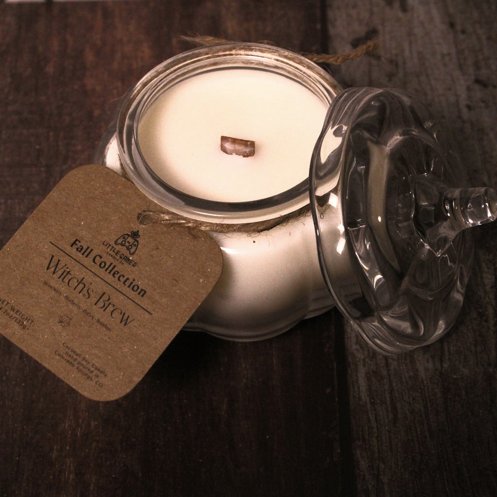 A glass jar candle with white wax and a small wooden wick, shown on a dark wooden surface with its lid leaning beside it and a kraft paper tag reading 'Fall Collection Witch Brew'.