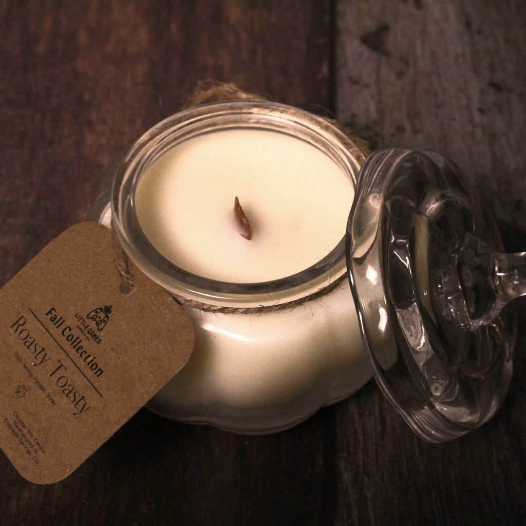 A single-wick candle in a clear glass jar with a matching glass lid resting beside it on a dark wooden surface. A kraft paper tag is attached to the jar and reads 'Roasty Toasty'.