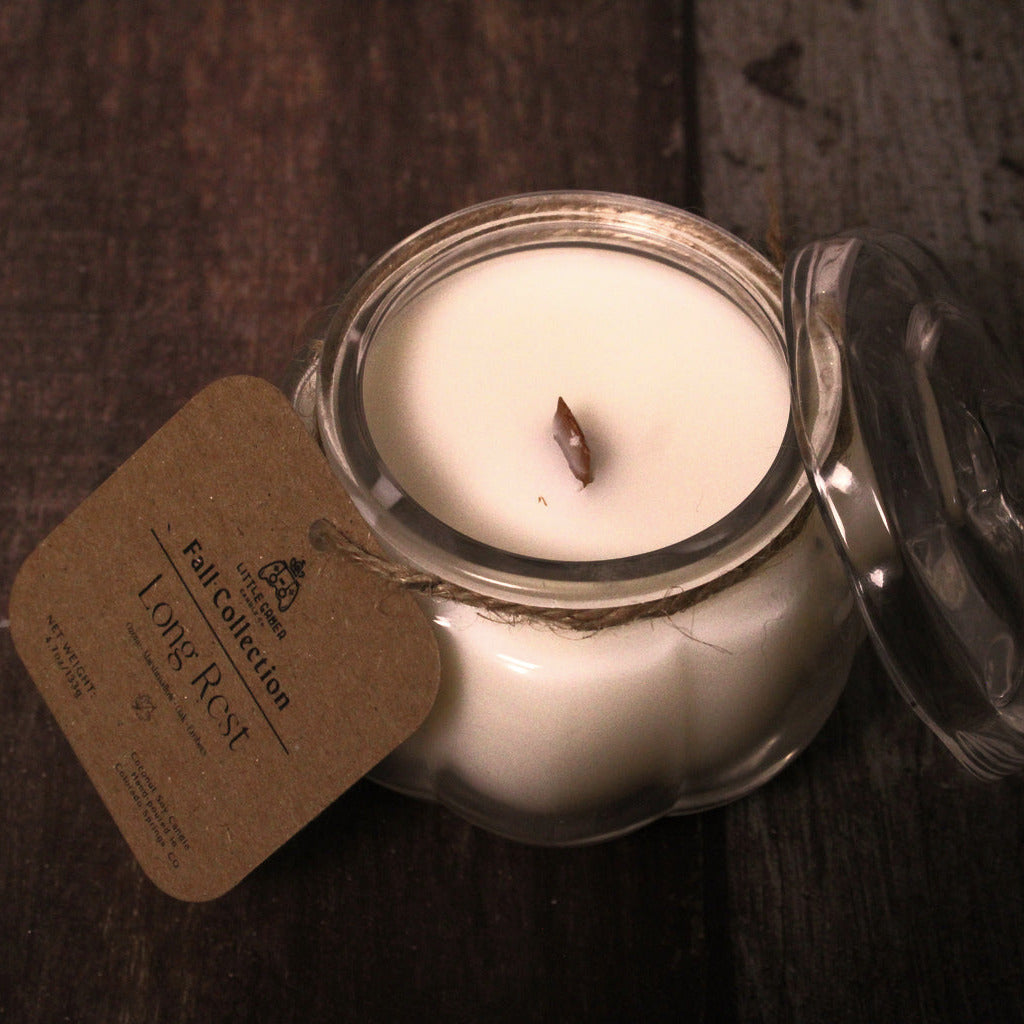 An open glass jar candle with white wax and a single wick, shown on a dark wooden surface with its glass lid leaning beside it and a kraft paper tag attached.