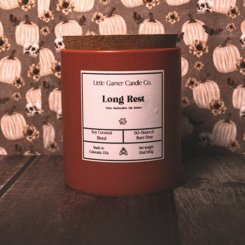 A red wax candle in a glass jar with a brown wooden lid, labeled 'Little Gamer Candle Co. Long Rest', sitting on a dark wooden surface in front of a pumpkin-patterned background.