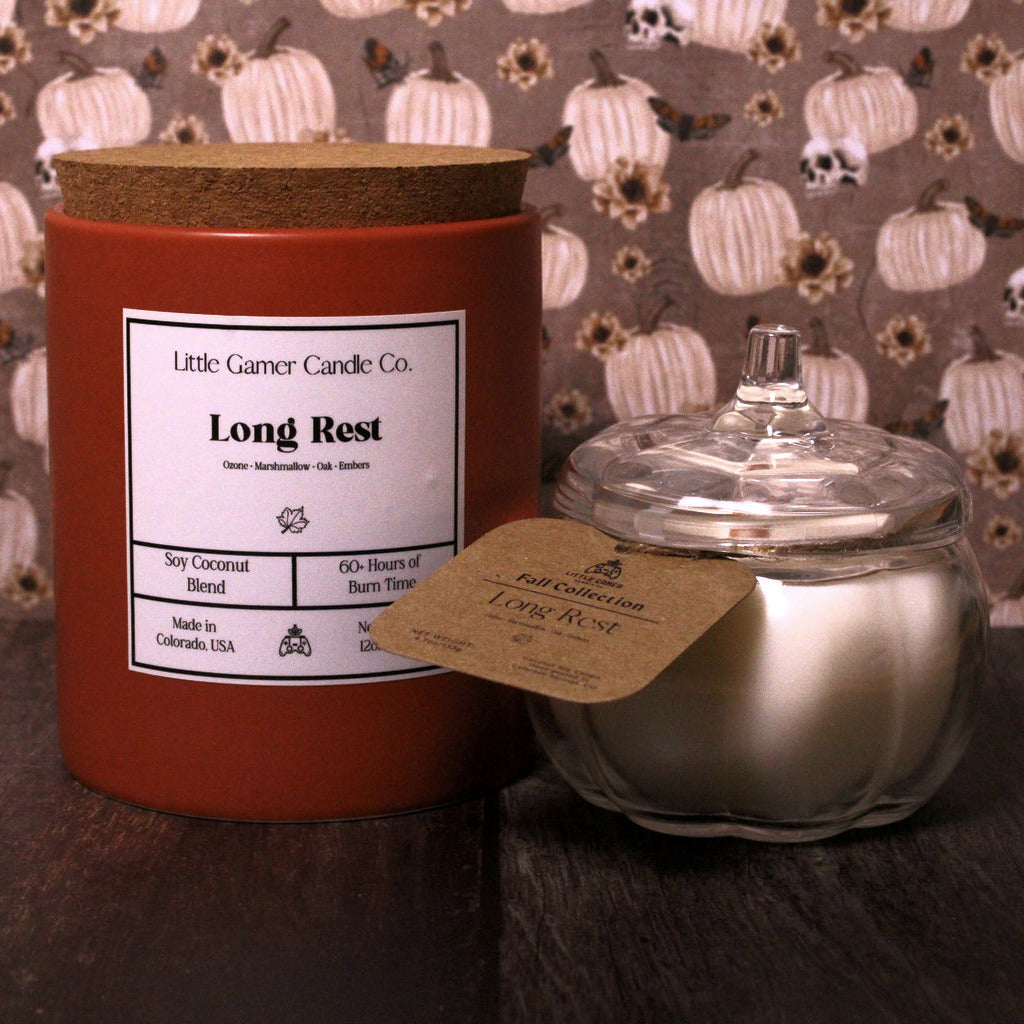 A small scented candle set on a dark wooden table, featuring a large terracotta-colored jar with a cork lid and a smaller silver glass jar with a clear glass lid. Both jars have kraft paper labels, with the larger jar's label reading 'Little Gamer Candle Co.