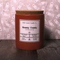 A small red wax candle in a cylindrical jar with a cork lid and a white label reading 'Little Garner Candle Co. Roasty Toasty,' set on a dark wooden surface in front of a pink ghost-patterned backdrop.