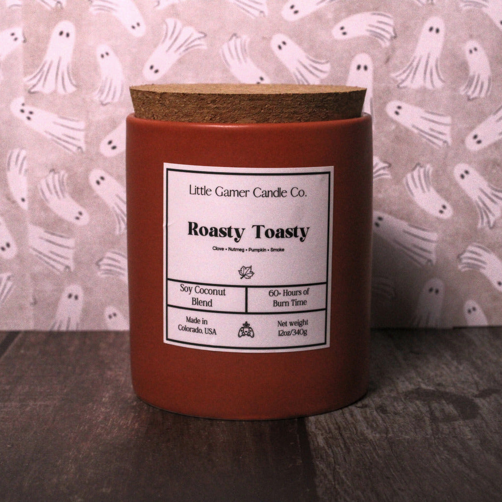 A small red wax candle in a cylindrical jar with a cork lid and a white label reading 'Little Garner Candle Co. Roasty Toasty,' set on a dark wooden surface in front of a pink ghost-patterned backdrop.
