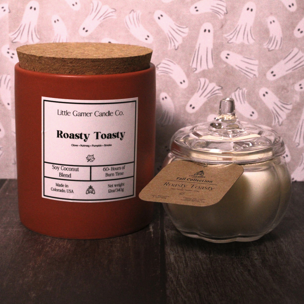 A red wax candle in a cylindrical jar with a cork lid stands next to a small clear glass jar containing metallic gold wax and a kraft paper tag. The background is a pink patterned sheet with ghost motifs, and the products are placed on a dark wooden surface.